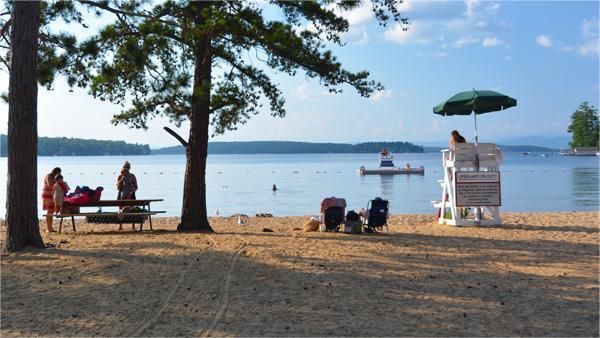 Sandy beach with people near water. Lifeguard on duty, picnic table, trees, and lake in the background.