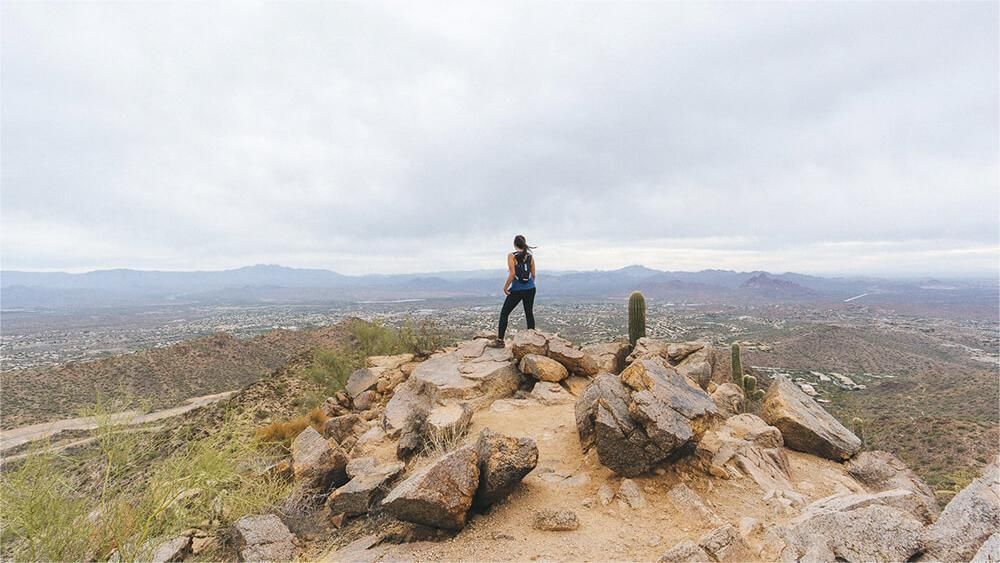 Woman standing on a rocky mountain, looking over a city under cloudy sky.
