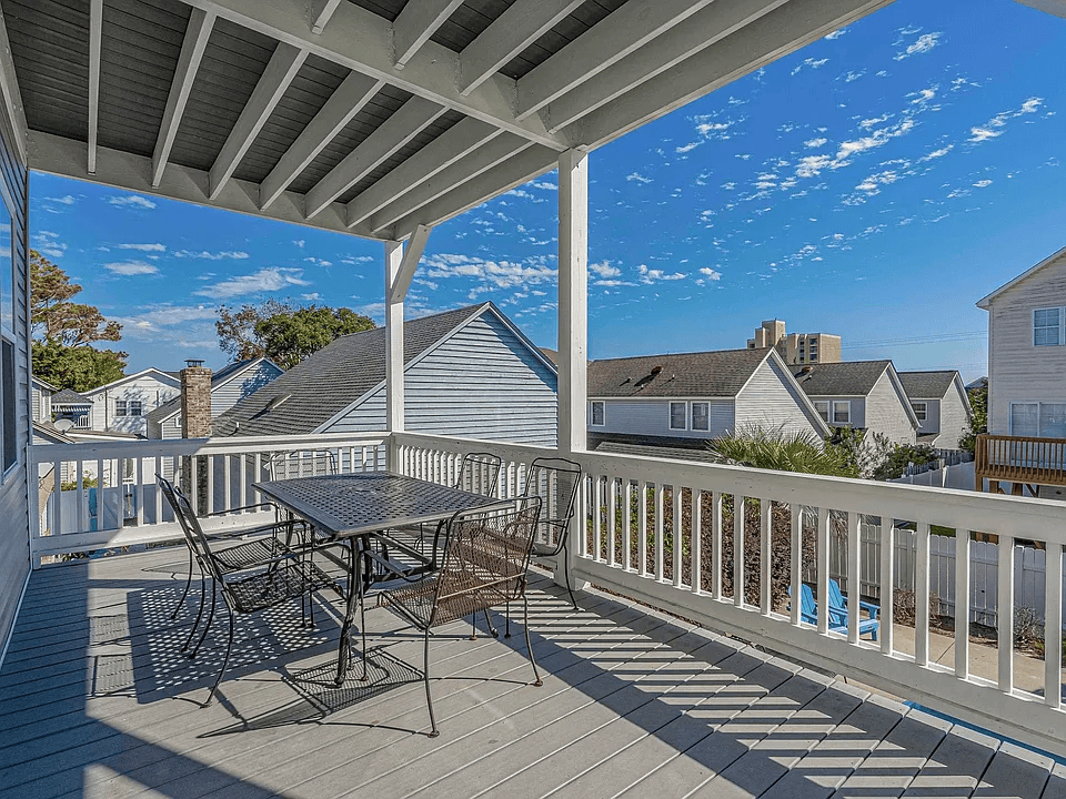 Covered porch with metal table and chairs, white railing, and a blue sky over neighboring houses.
