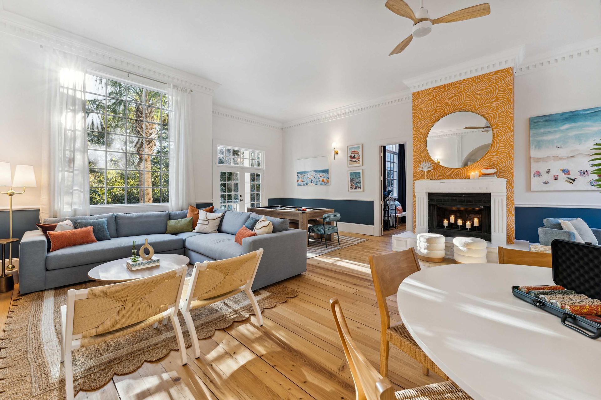 Bright living room with gray sofas, white chairs, a fireplace, large windows, and an orange accent wall
