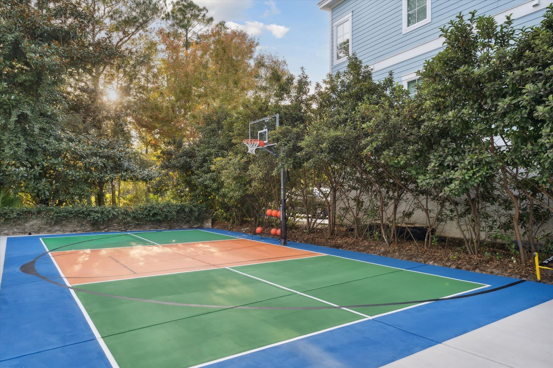Outdoor backyard basketball half-court with blue, green, and orange surface beside trees and a house.