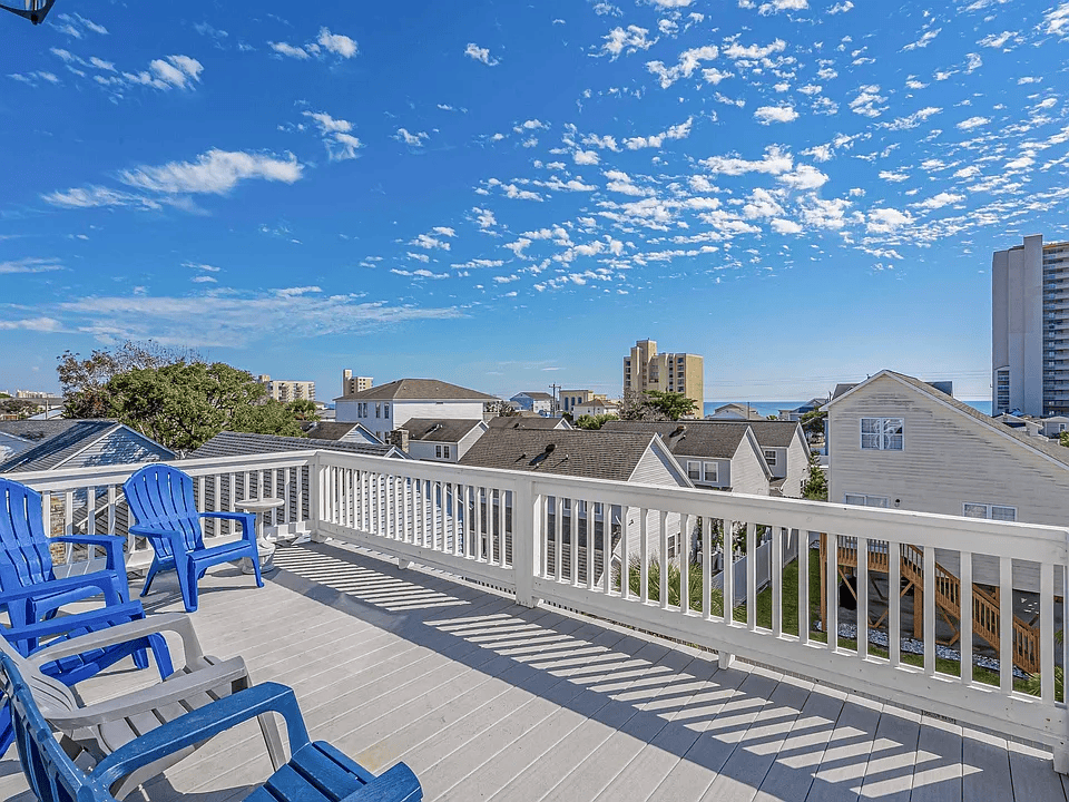 Blue chairs on a rooftop deck with white railing, overlooking houses and a city skyline under a bright blue sky.