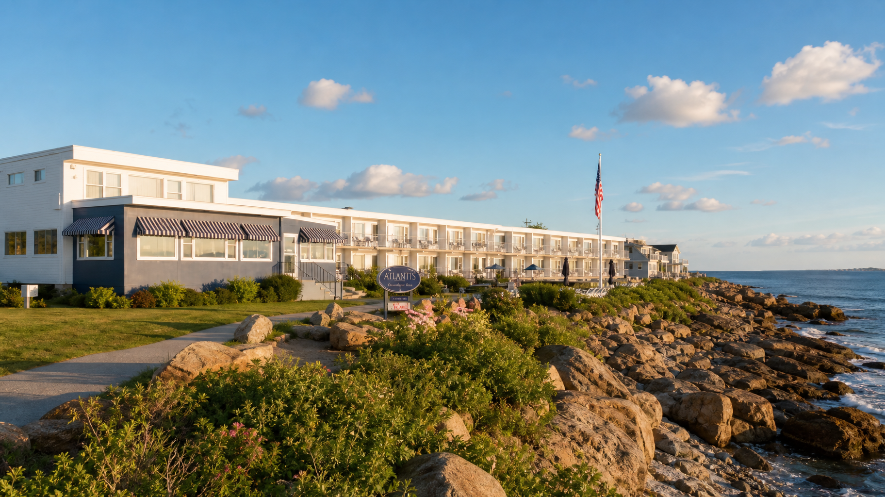Coastal hotel building beside rocky shoreline under a blue sky, with an American flag flying nearby