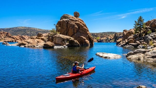 Person kayaking on a blue lake beside tan rock formations under a clear sky