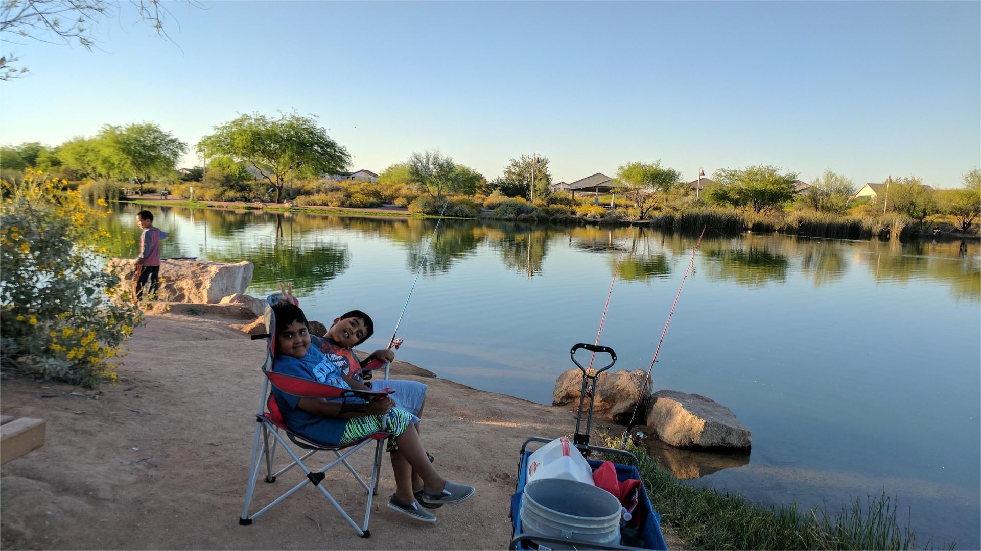 Two people fishing by a lake; one person seated in a chair, sunny day.