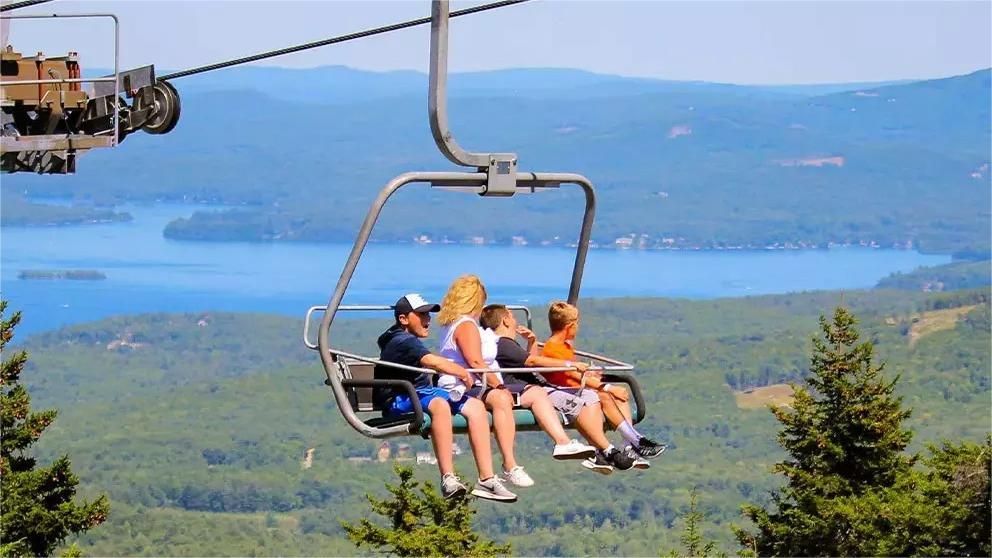 People on a ski lift overlooking a blue lake and forested hills.