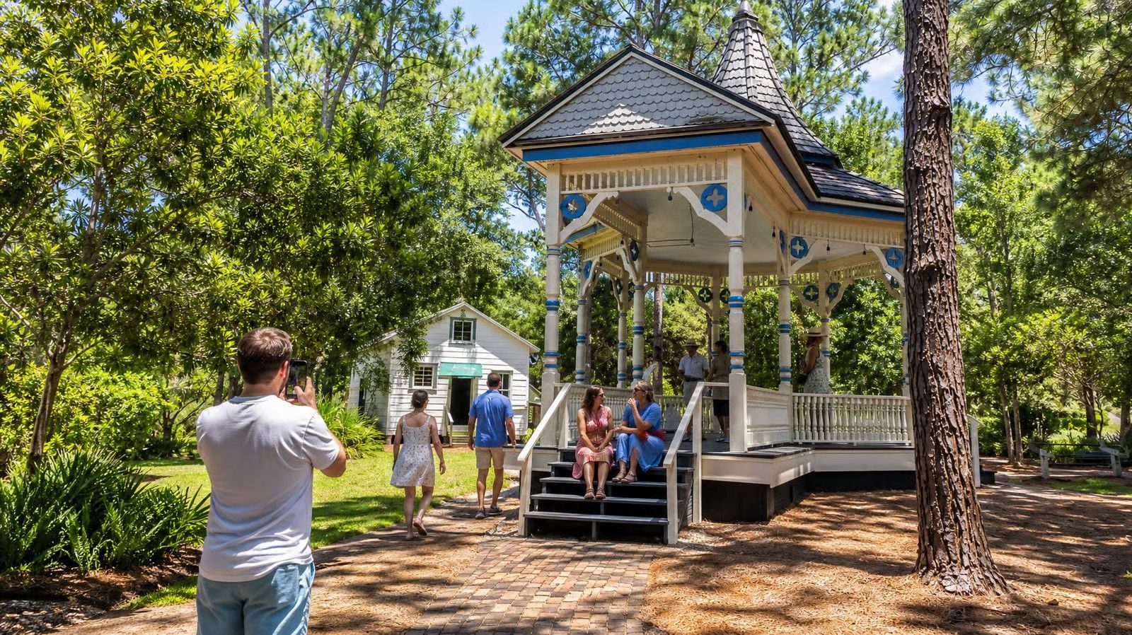 People gathered around a small white gazebo in a wooded park on a sunny day.