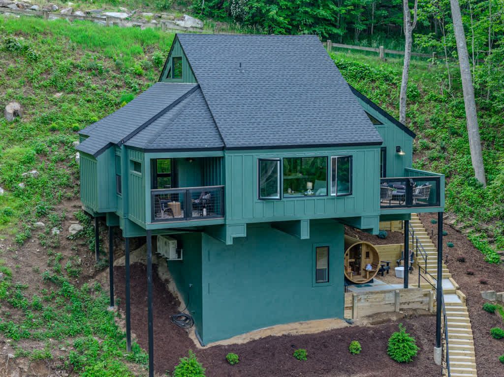 Small teal cabin with dark roof on a hillside, surrounded by greenery and a stone path