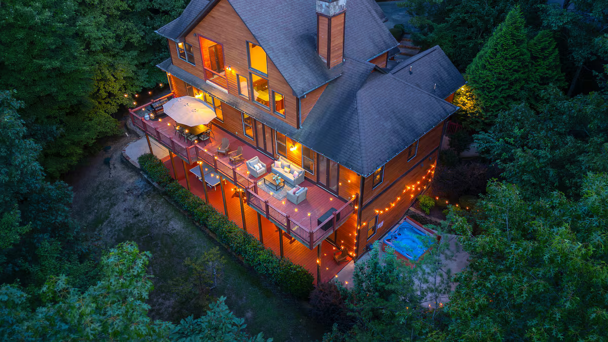 A warmly lit cabin with a wraparound deck, surrounded by dense green trees at dusk.