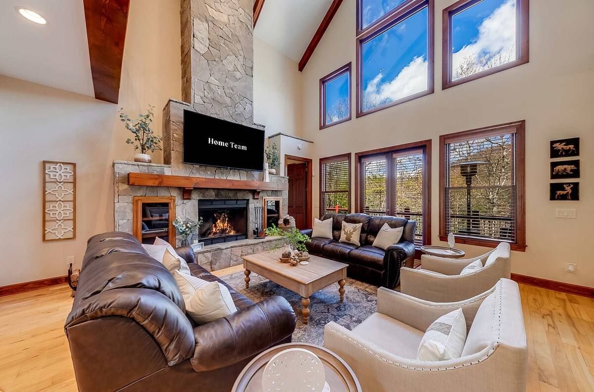 Spacious living room with leather sofas, stone fireplace, wall-mounted TV, and large windows overlooking trees