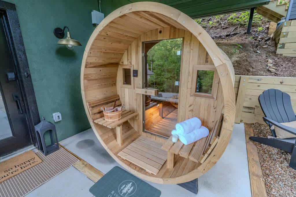 Wooden barrel sauna on a patio with a glass door, benches, and a small towel basket