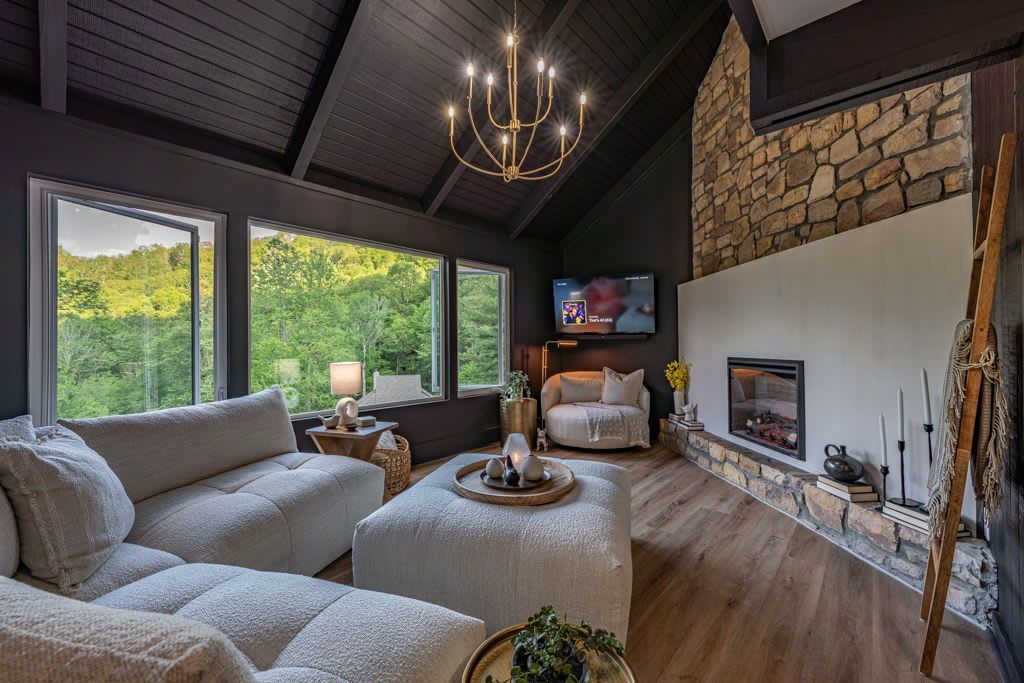 Cozy living room with gray sectional, round ottoman, stone fireplace, and large windows overlooking greenery