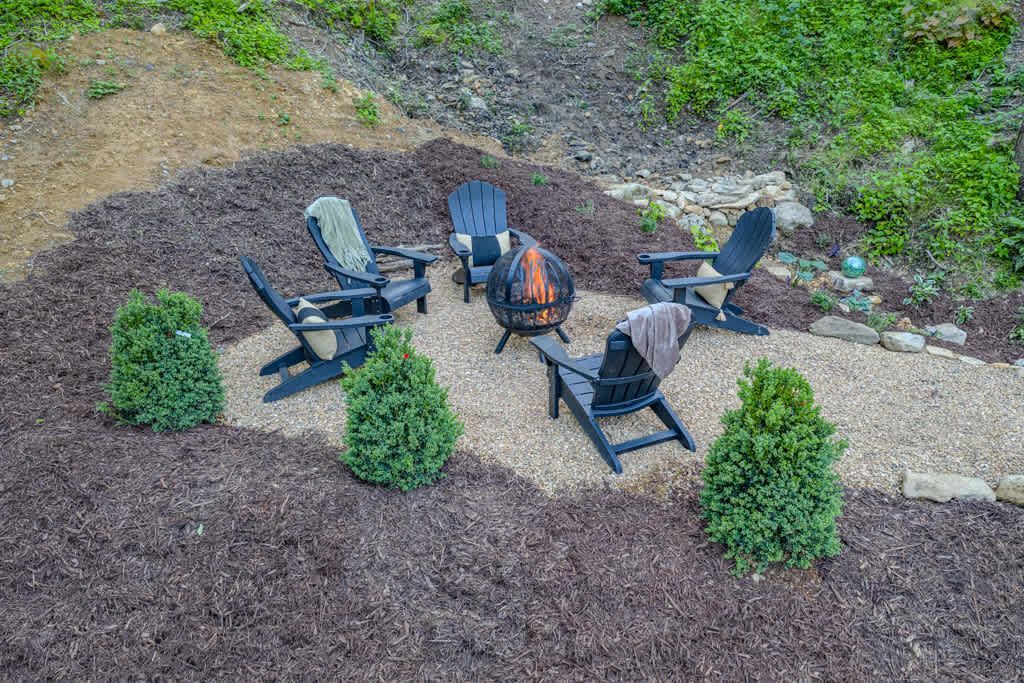 Four Adirondack chairs around a small fire pit in a landscaped yard with mulch and shrubs