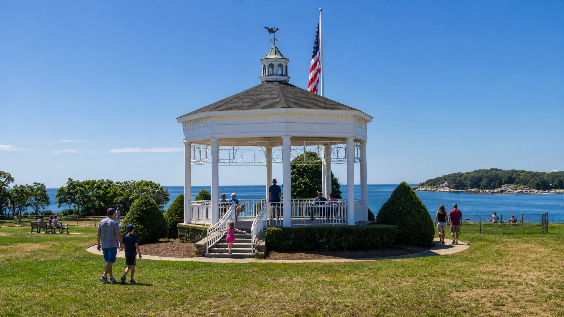 White gazebo by the water with an American flag, people nearby, and a sunny blue sky