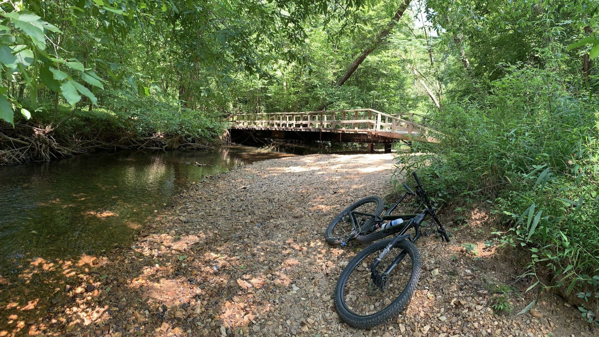 A mountain bike sits on a gravel riverbank near a small wooden bridge in a dense, green forest.