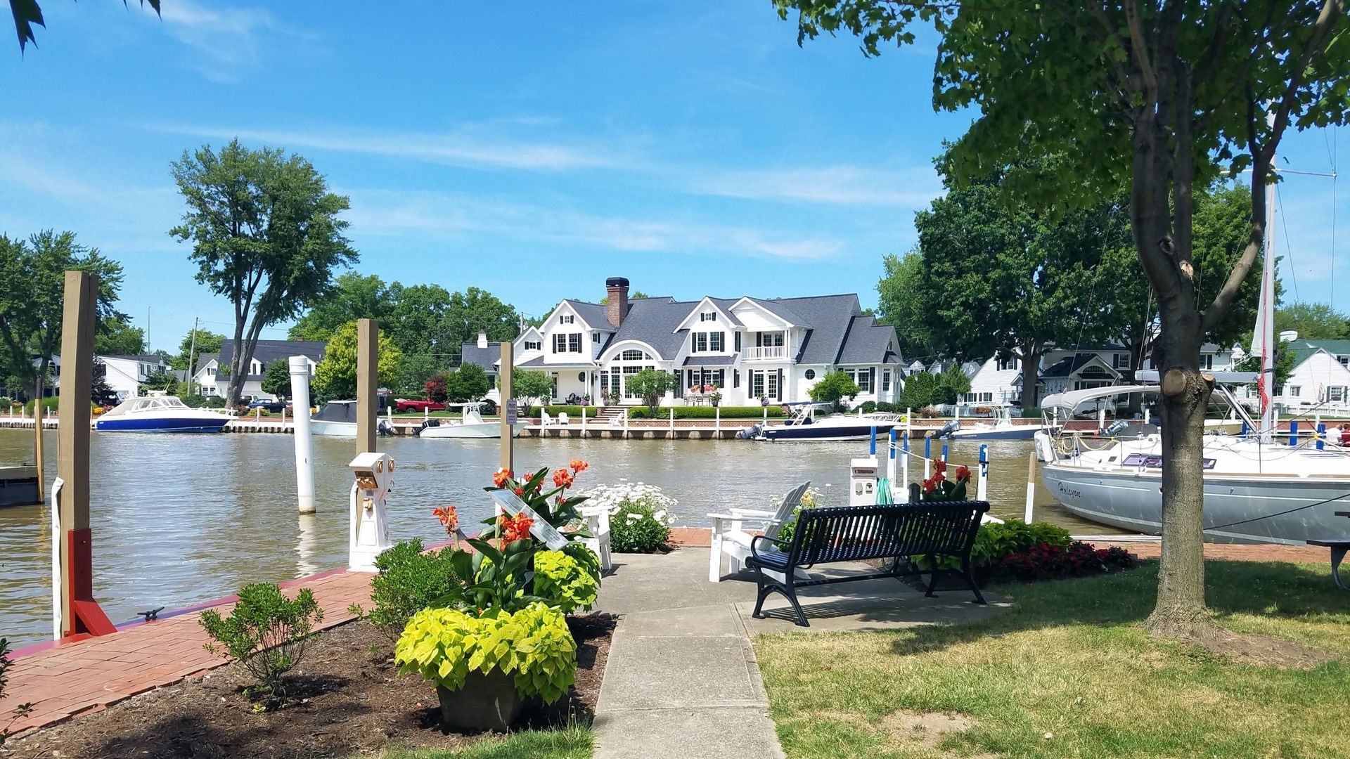Waterfront view with boats, houses, a walkway, trees, and flowers under a blue sky.
