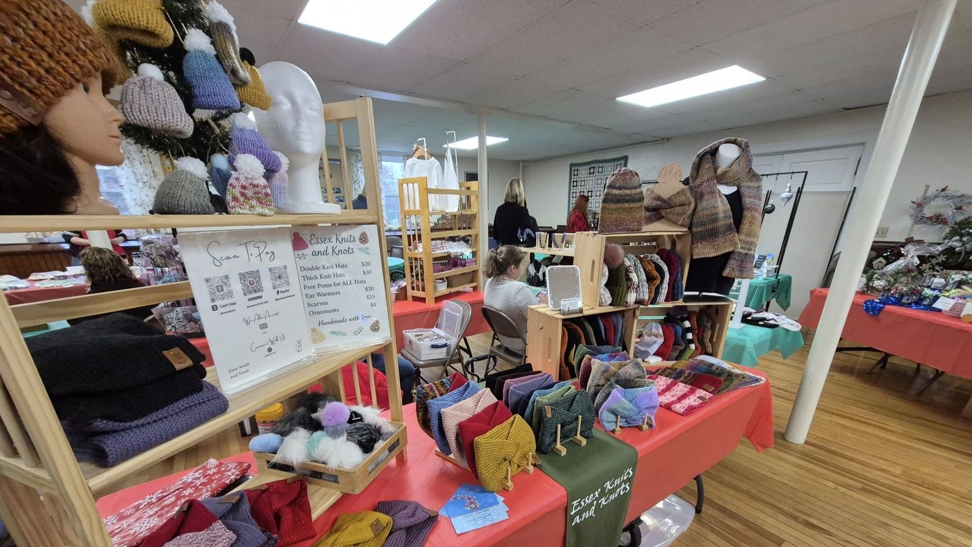 Inside a craft fair booth, hats and scarves are displayed on tables and shelves. A person is sitting at a table.