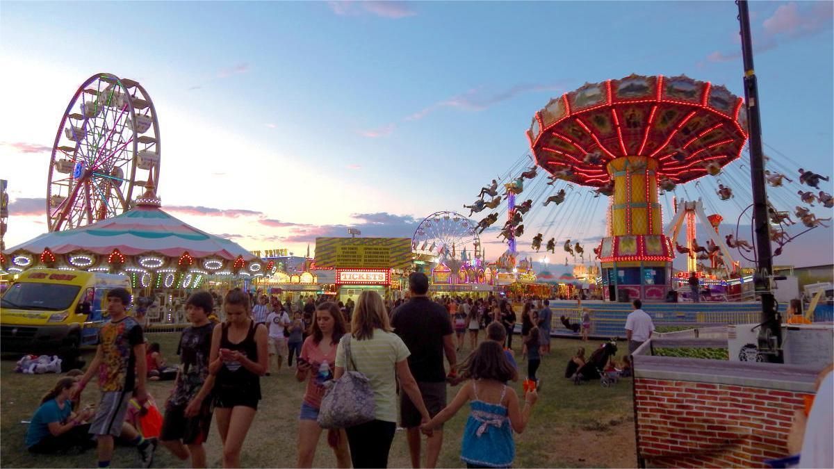 Carnival with Ferris wheel and swing ride at dusk; crowds of people walk about.