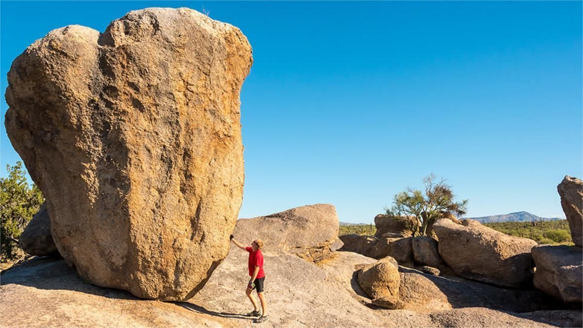 Person touching a large, balancing boulder in a desert landscape under a clear blue sky.