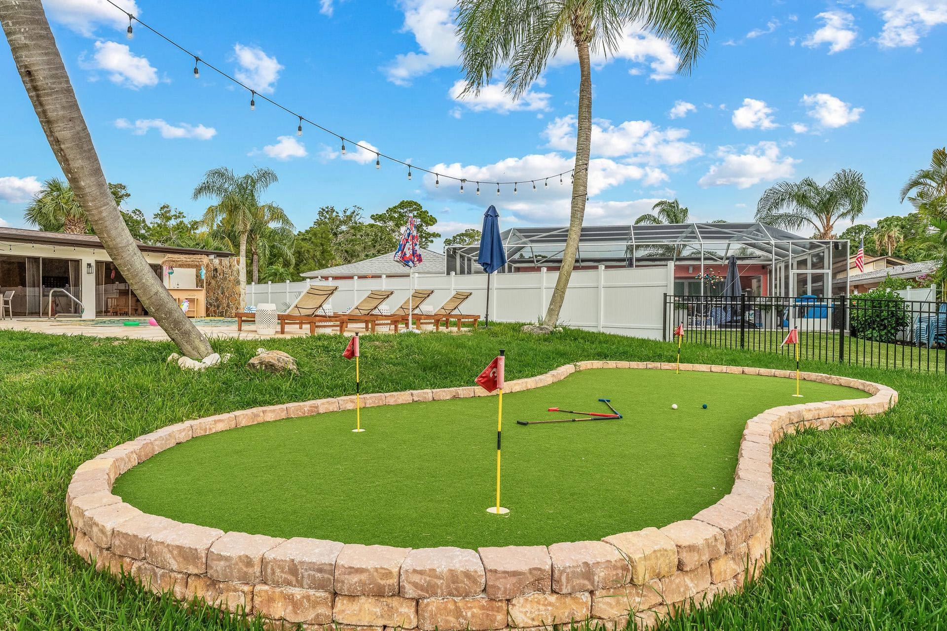 Mini golf green with a flagstick, stone border, and string lights in an outdoor park setting