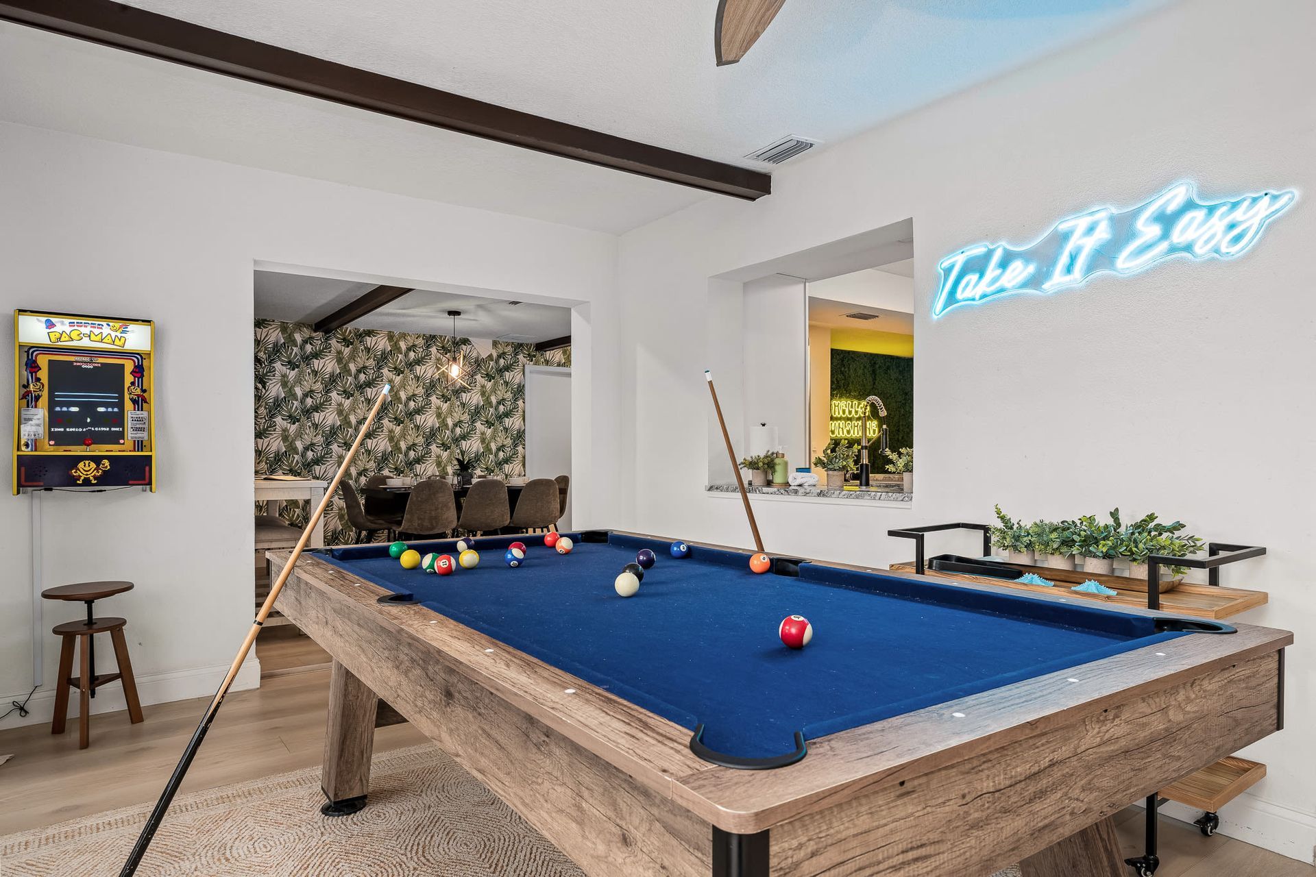 Pool table in a bright game room with neon sign and stools, blue felt and balls scattered on the table