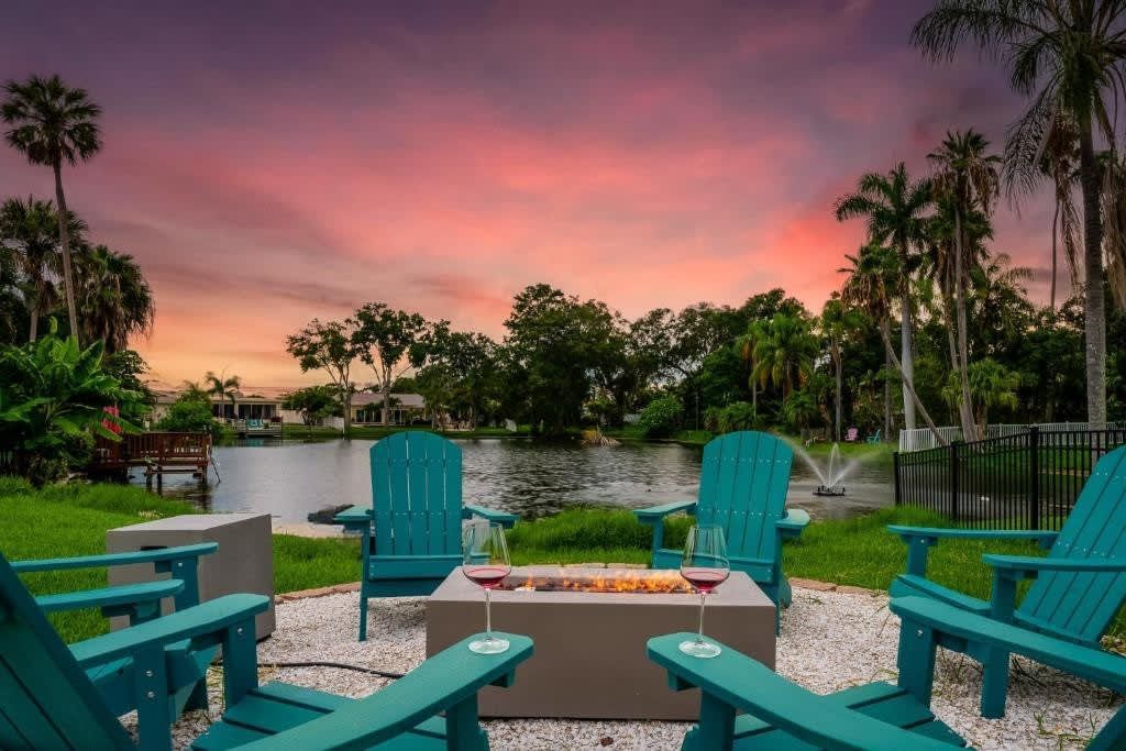 Teal chairs around a fire pit by a lakeside at sunset, with palm trees and pink sky