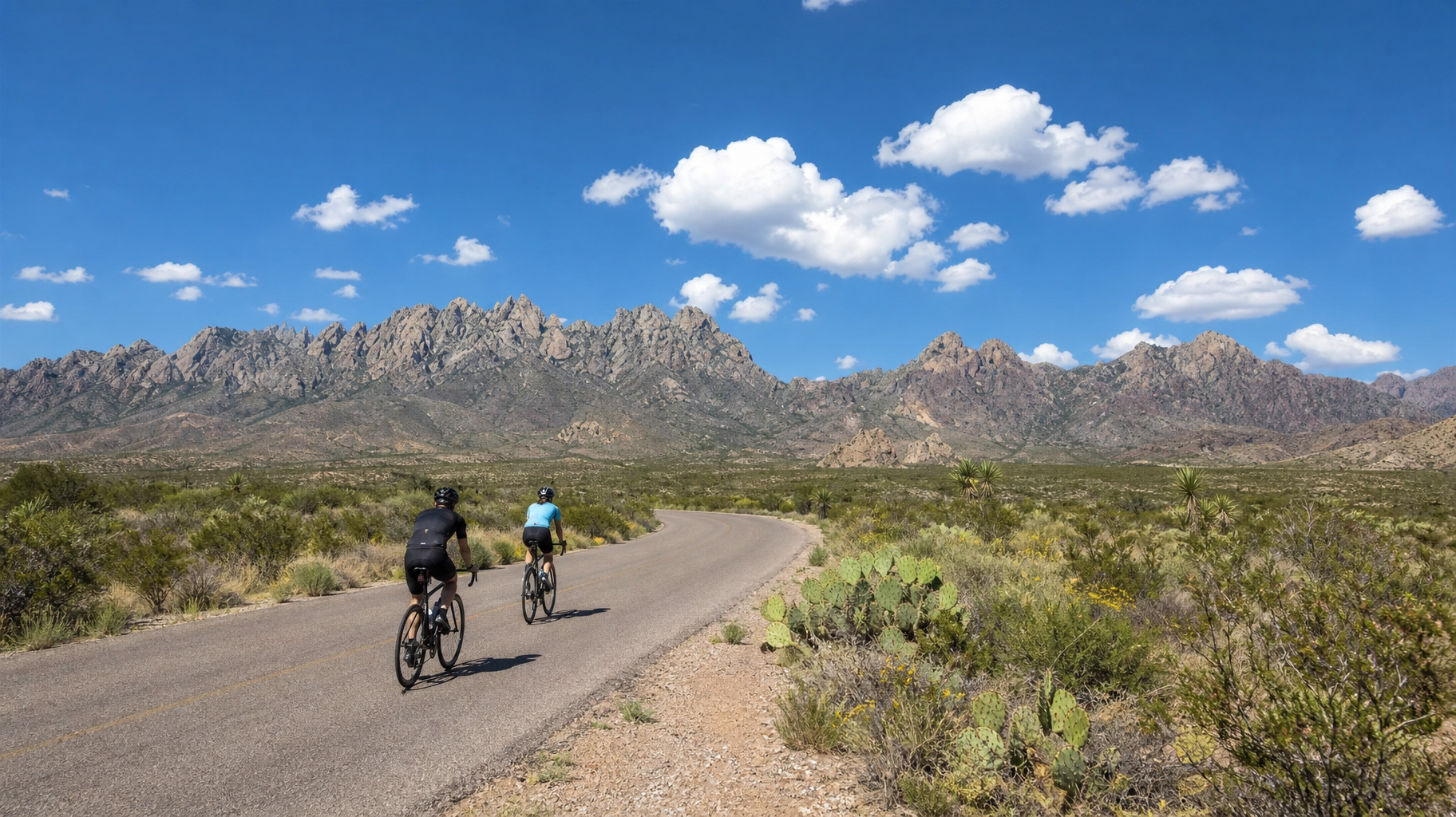 Two cyclists ride on a desert road with mountains, shrubs, and a bright blue sky with clouds.
