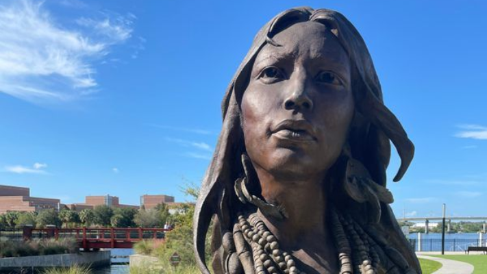 Bronze statue of a woman with long hair, looking forward, against a blue sky with buildings and water in the background.