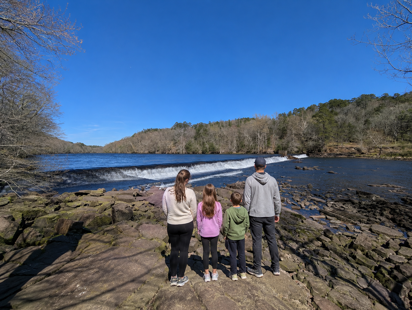 A family of four stand on rocky riverbank by a wide river under a bright blue sky at the Beavers Bend State Park in Oklahoma