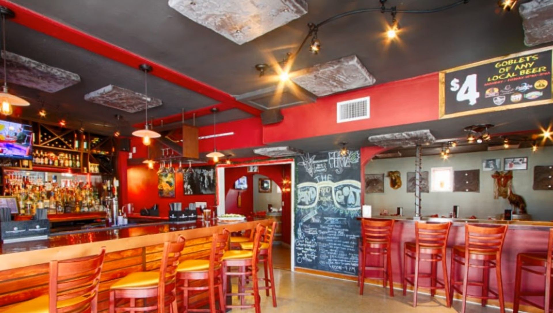 Interior of a bar with red accents, wooden bar, stools, and chalkboard menu.