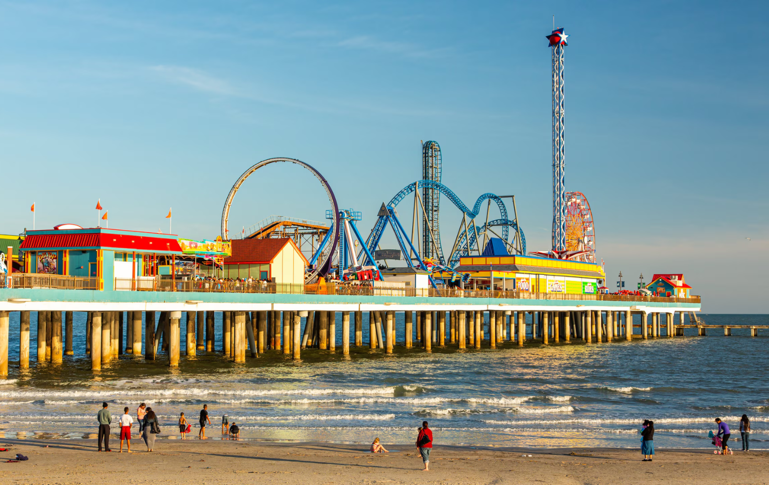 People are walking on the beach near a pier with a roller coaster in the background in Galveston, Texas