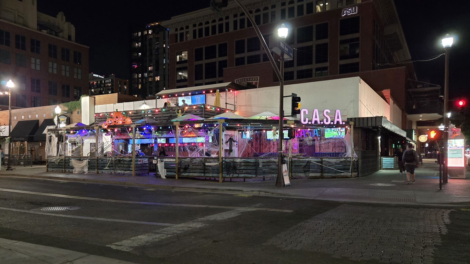 Nighttime view of a brightly lit bar called CASA with outdoor seating. Pedestrians walk on the sidewalk.
