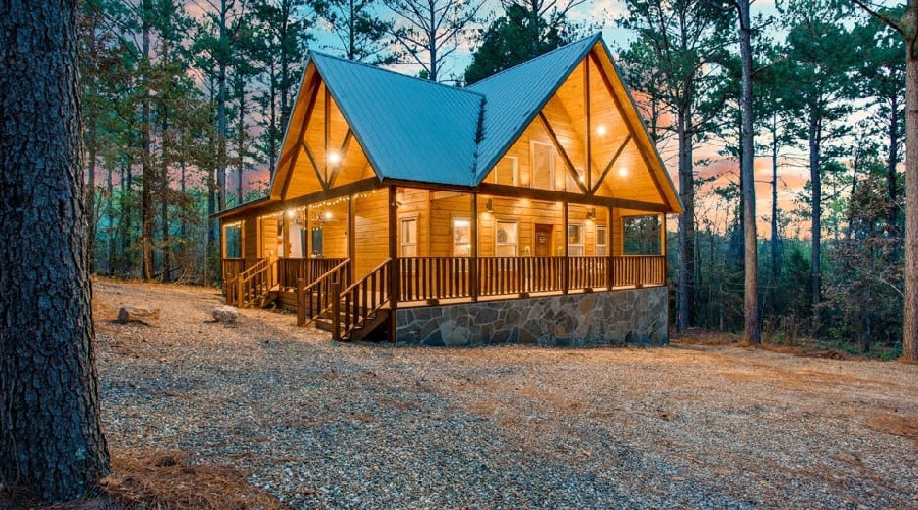 Cabin in the woods with lit windows and a wraparound porch, surrounded by trees and gravel.