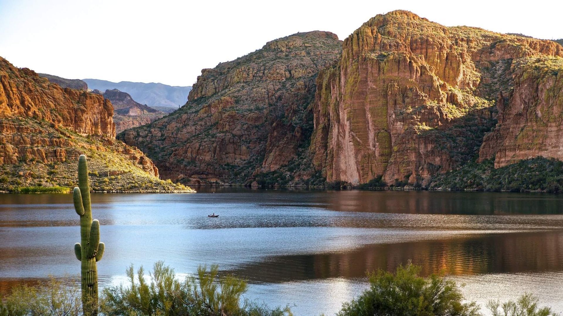 Lake surrounded by rocky, mountainous terrain and desert vegetation, with a cactus in the foreground.