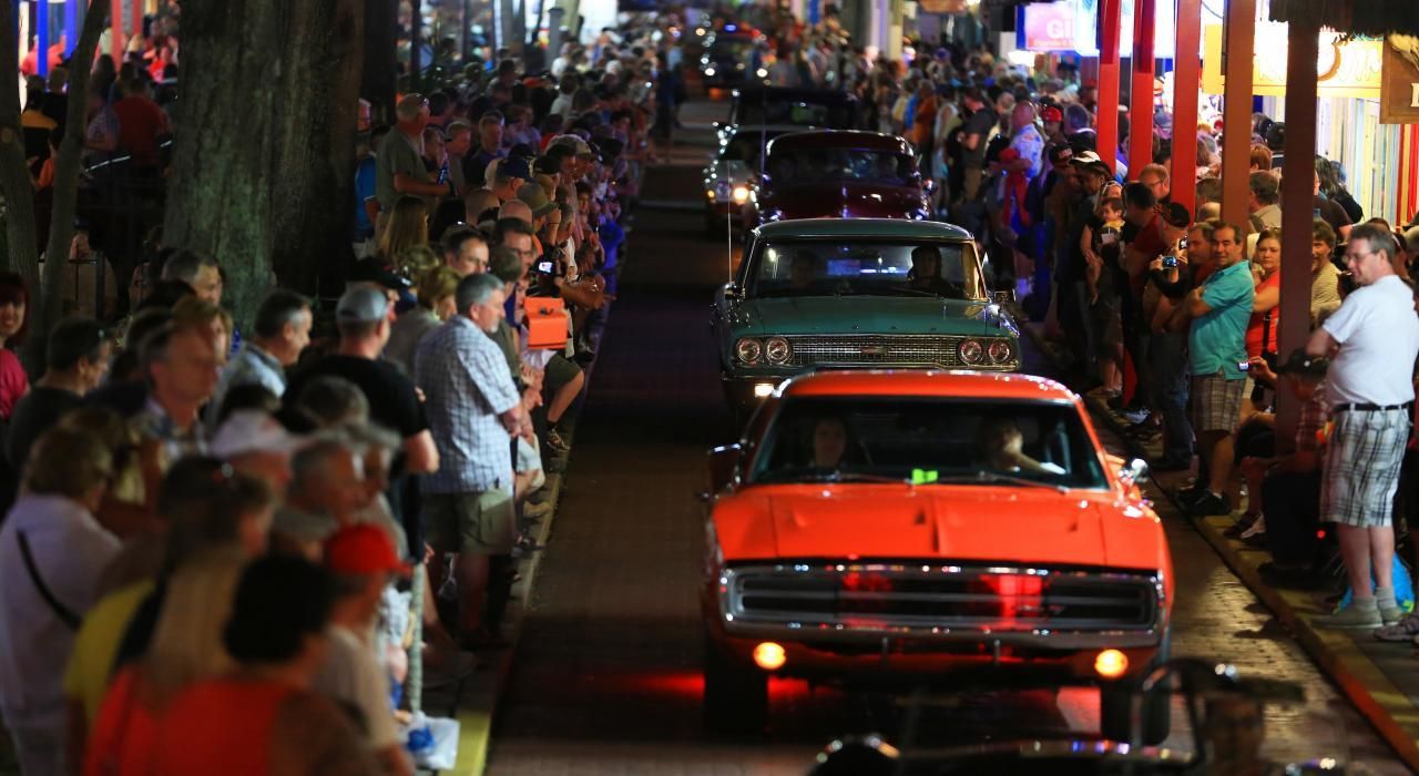 Classic cars driving down a street lined with spectators at night. Orange car leads, green truck follows.