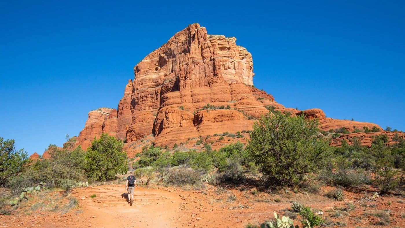 Person hiking towards a large red rock formation under a bright blue sky.