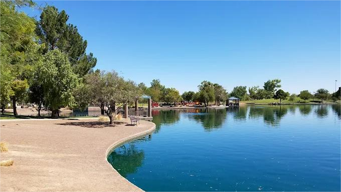 Lakeside park on a sunny day. Blue water reflects trees and sky. Sandy shore in foreground.