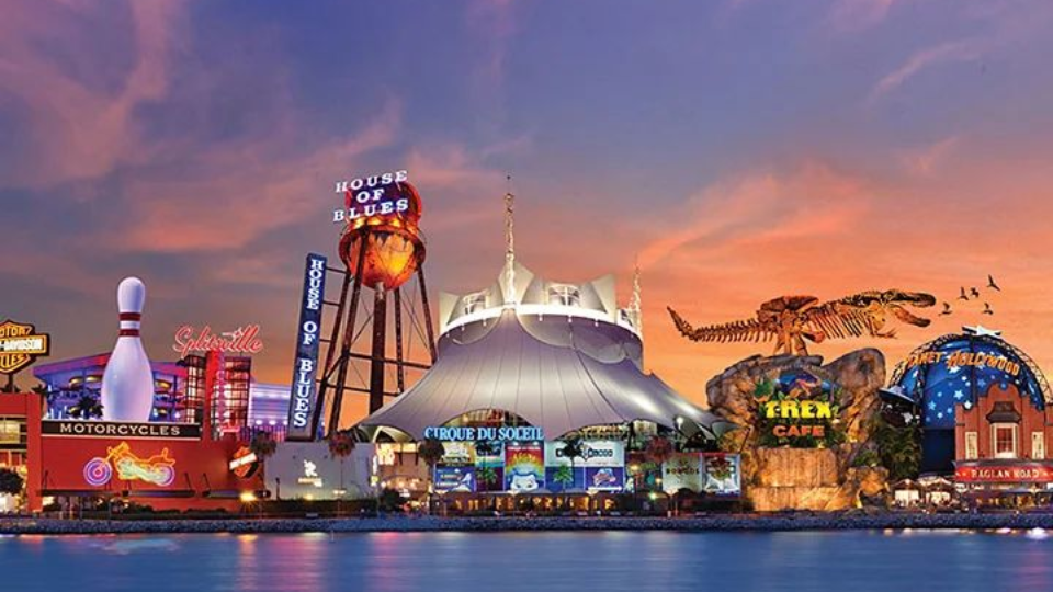 Skyline of Disney Springs at sunset, with neon signs, a water tower, and large buildings over water.