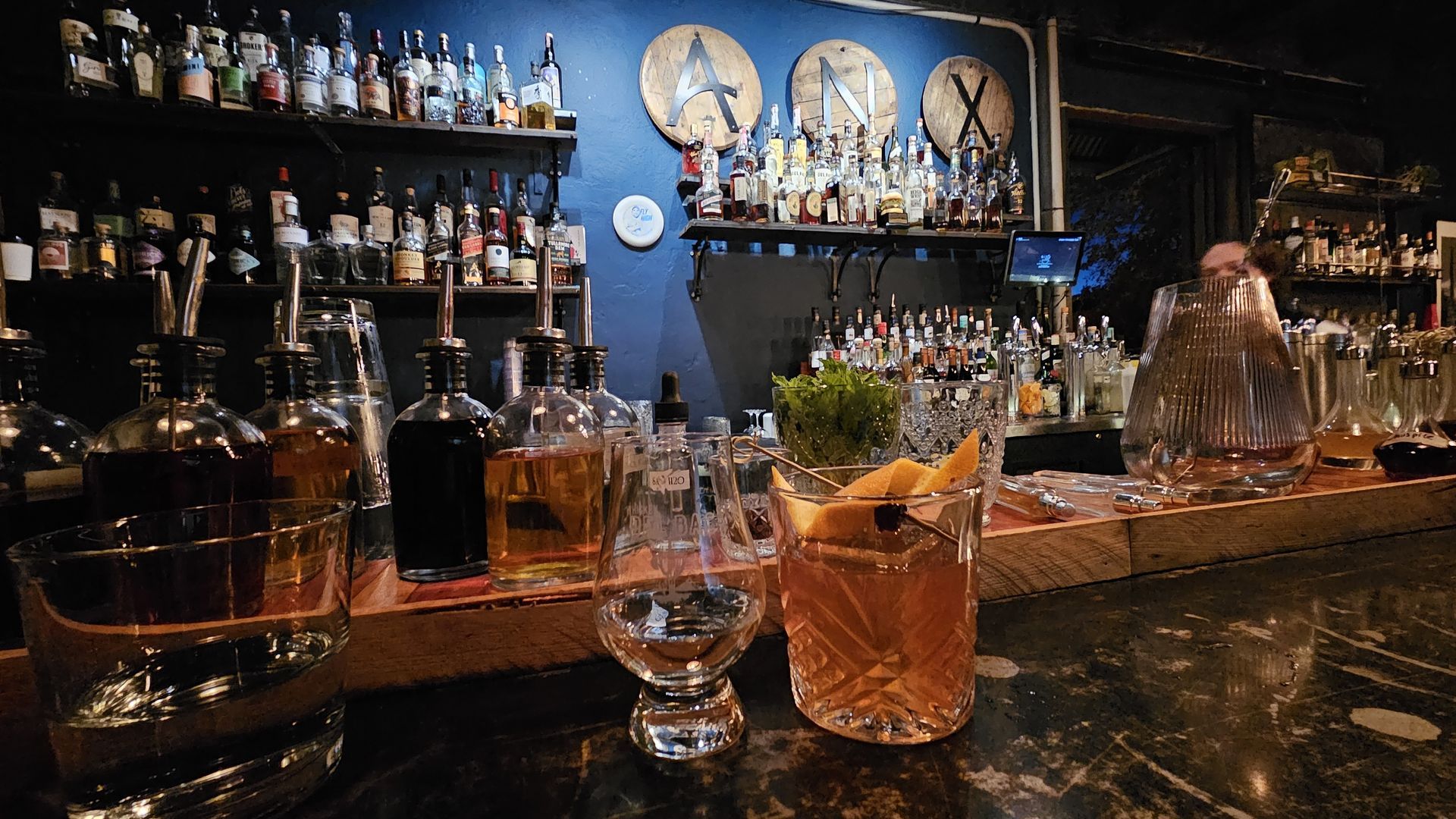 Cocktail bar with bottles on shelves, drink on counter, garnished with orange peel and mint.