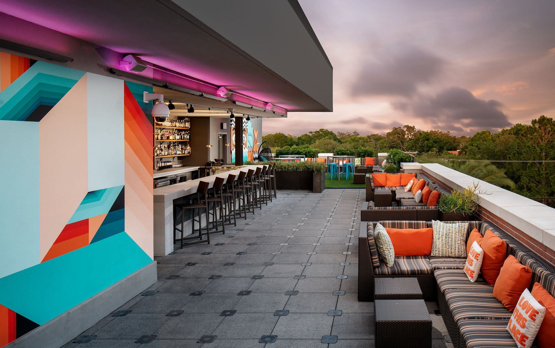 Rooftop bar with seating, stools, and colorful mural. Cloudy sky above trees.