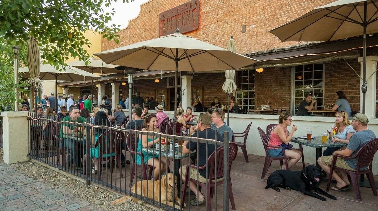 Outdoor café patio with seated diners under umbrellas beside a brick building and iron fence