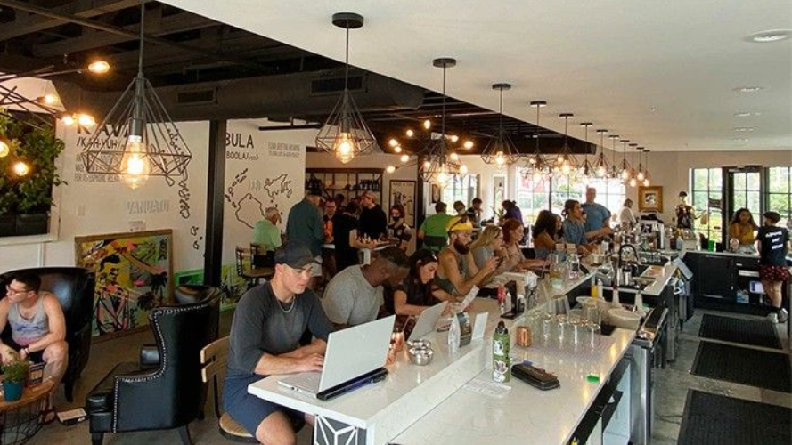 Bar scene, people at counter, some with laptops. Overhead lights, art on wall, open to the outdoors.
