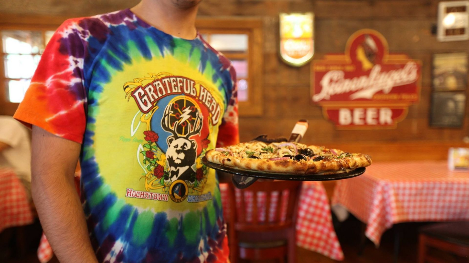 Server holding pizza, wearing tie-dye Grateful Dead shirt, in a restaurant with checkered tablecloths.