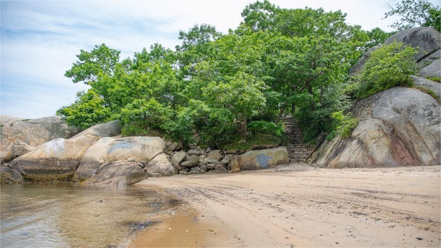 Sandy beach with large rocks, green trees, and blue sky.