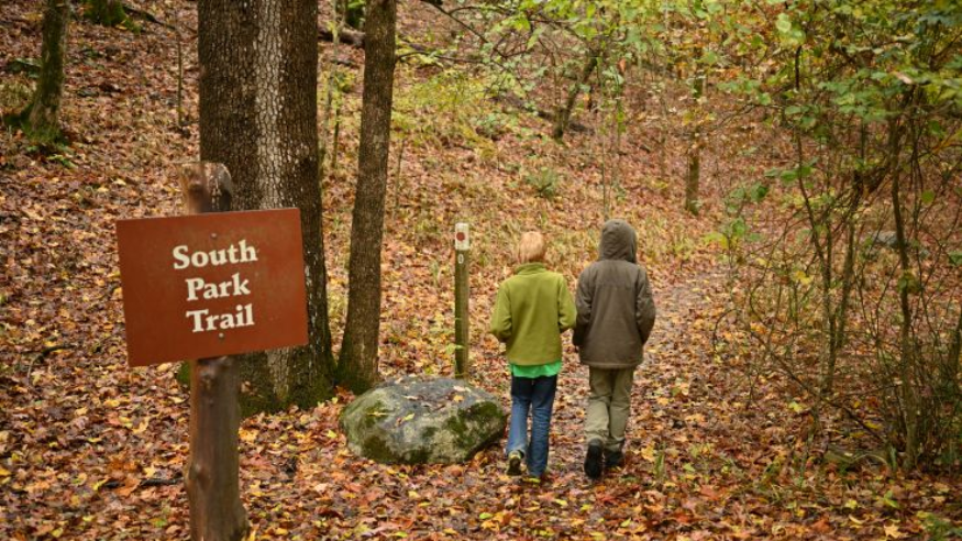 Two people walk on a leaf-covered trail in South Park. A sign marks the South Park Trail.