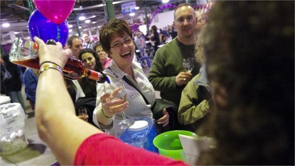 Woman laughing, being poured a drink at a tasting event, surrounded by other people. Balloons and blue bin visible.