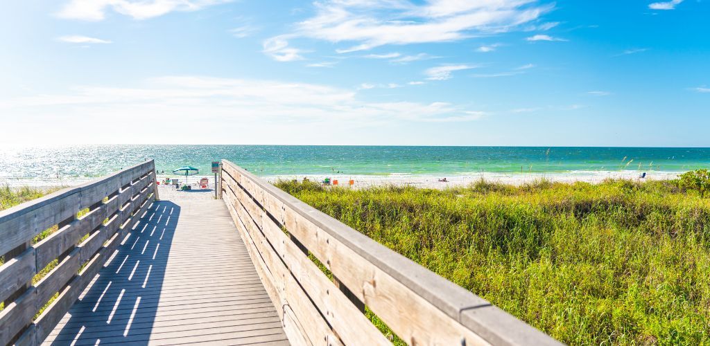 Wooden boardwalk leading to a sunny beach with green water and blue sky.