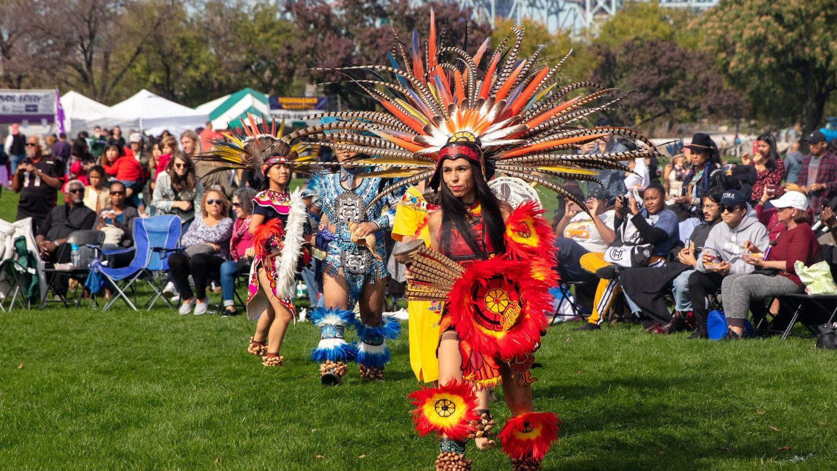 Performers in elaborate, colorful feather headdresses and traditional regalia dance on a grassy field before a crowd.