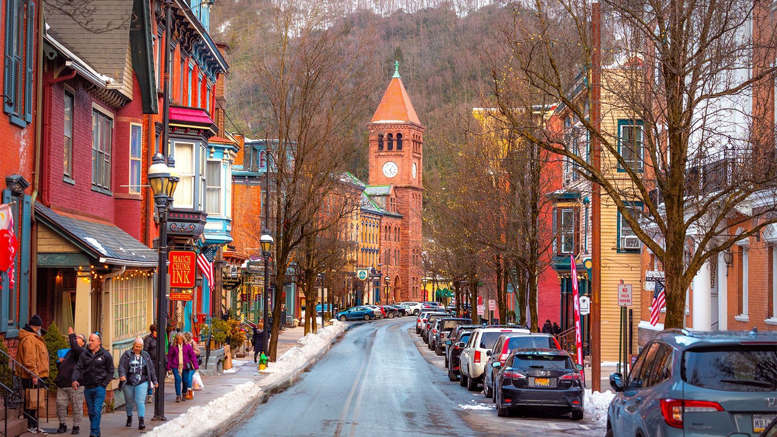 Street lined with colorful buildings and parked cars, toward a brick clock tower. People walk on sidewalks.