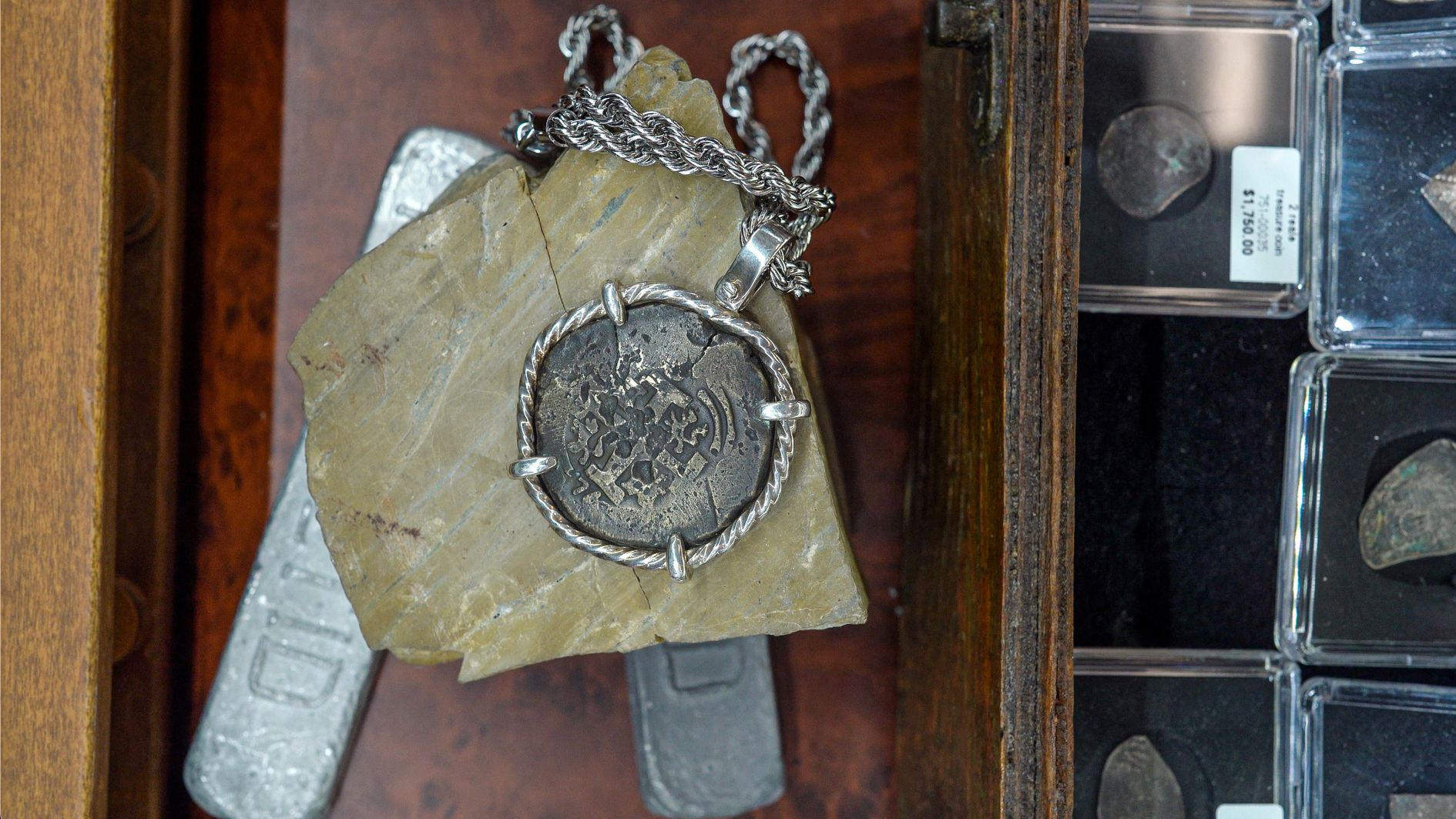 Close-up of a silver coin pendant on a chain, resting on a paper, in a wooden drawer next to silver bars and coin containers.